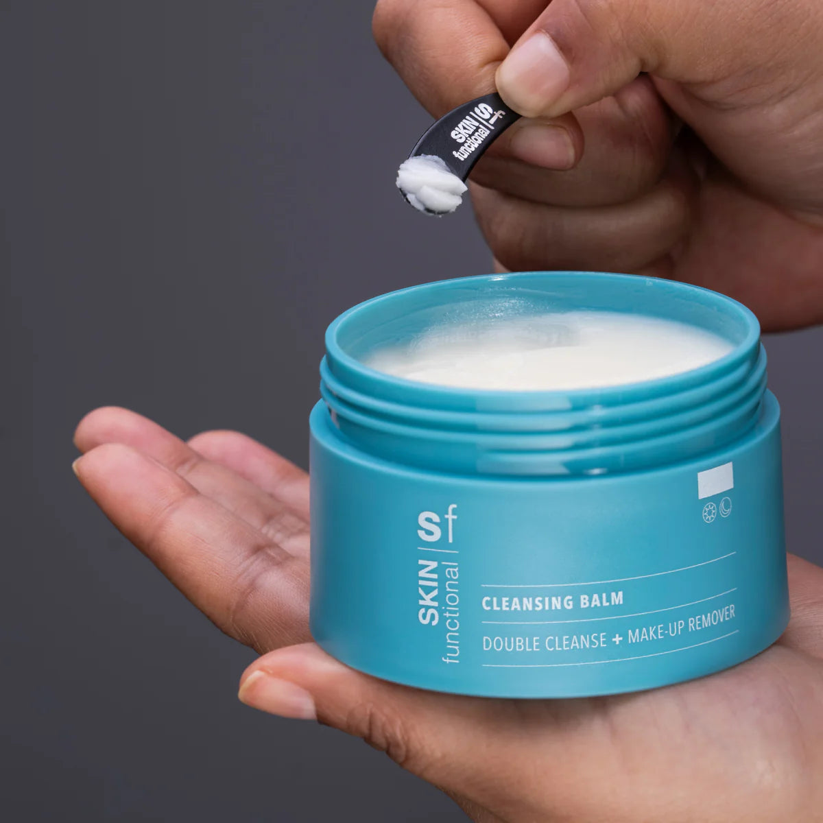 Blue jar of cleansing balm with a hand holding a applicator against a gray background